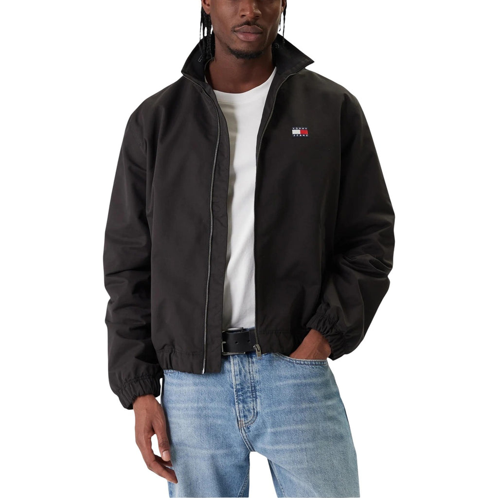 Detail close-up of the black blazer featuring a zipper closure and logo patch on a white background