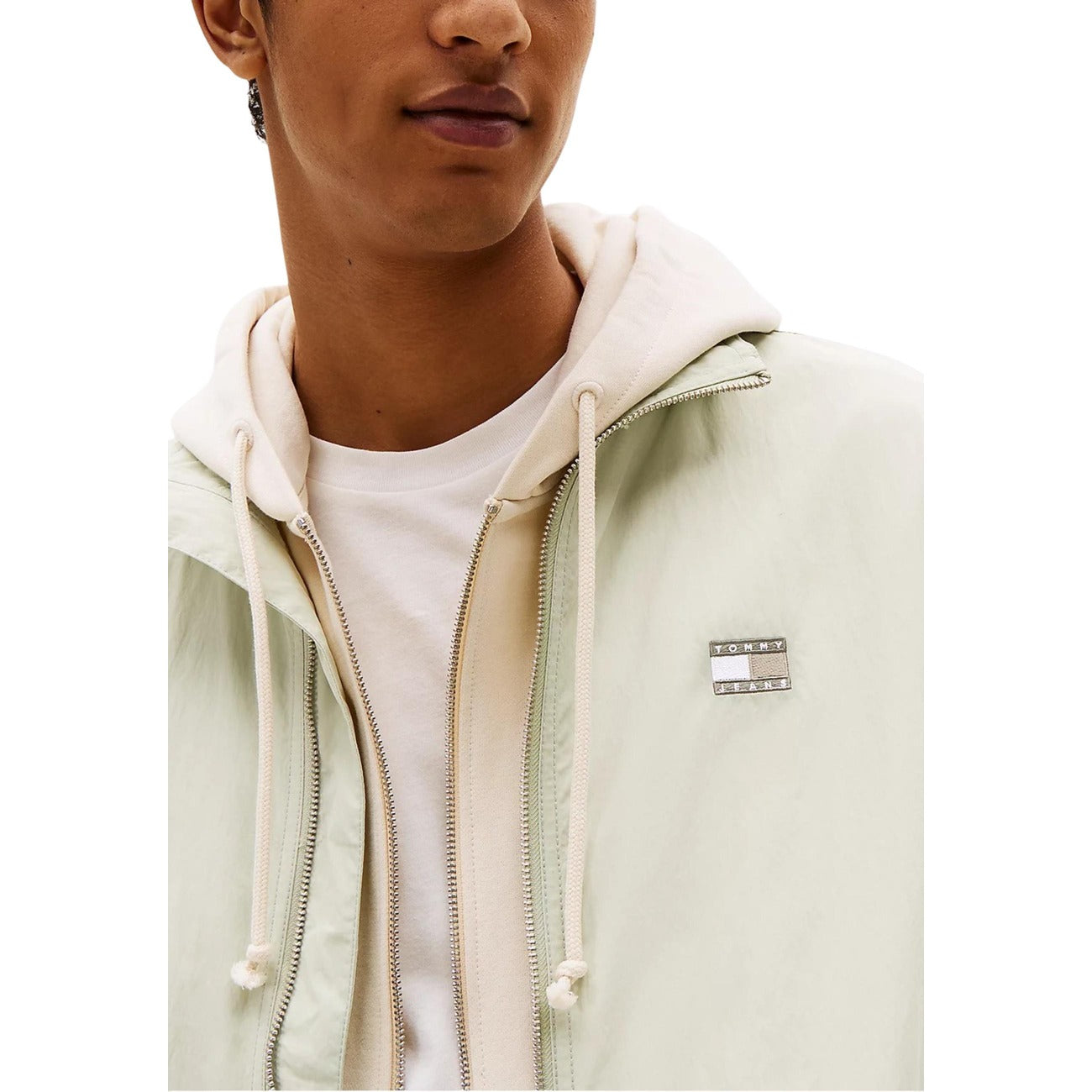 Detail close-up of a light green jacket featuring a zipper closure and logo patch on a white background